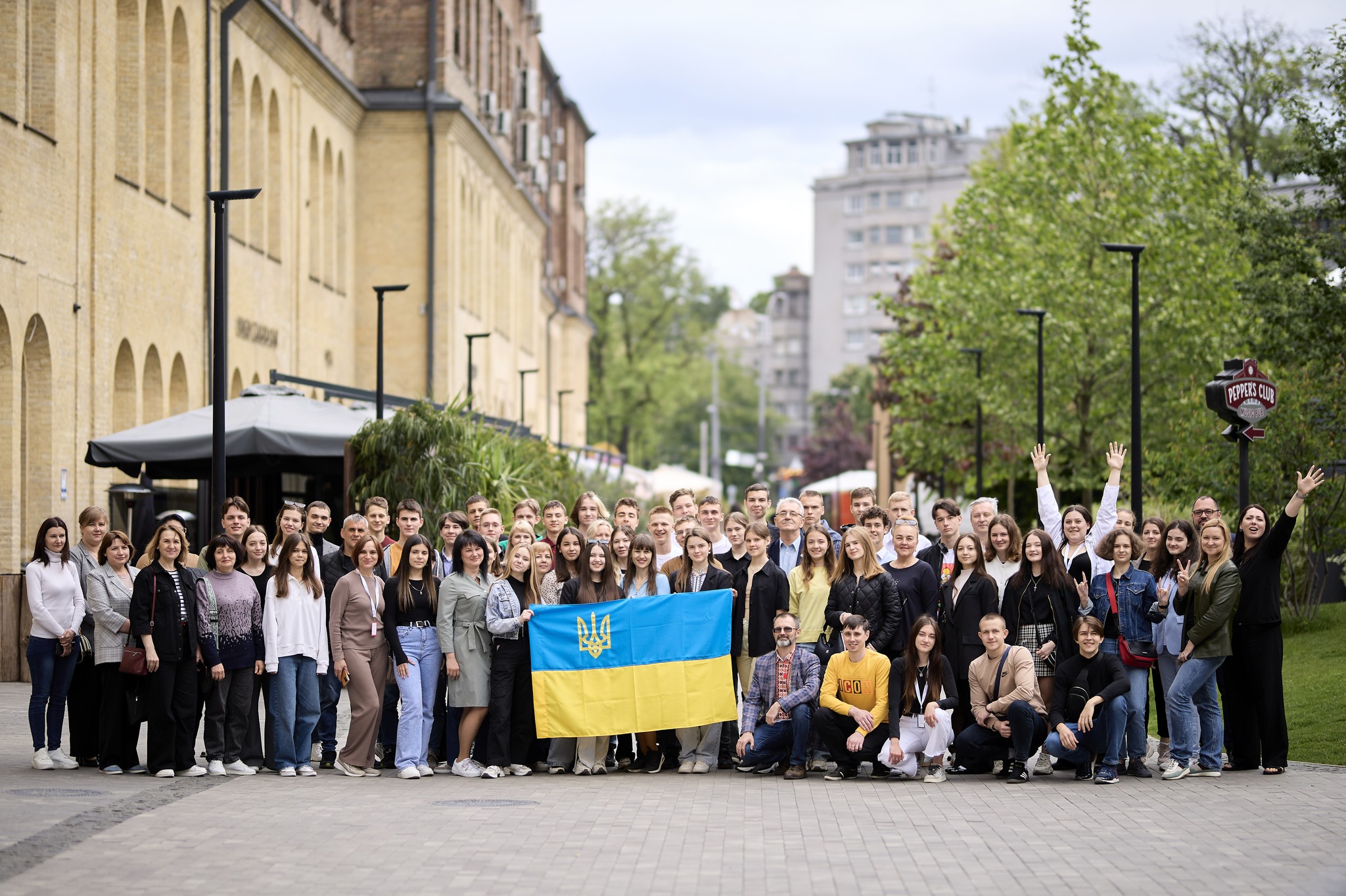A large group photo taken outside of adults and students, behind the Ukrainian flag being held up by two students